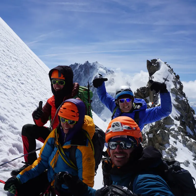 Vier bergbeklimmers met helmen, zonnebrillen en winterkleding glimlachen en poseren op een besneeuwde berghelling, met rotsachtige toppen en een blauwe lucht op de achtergrond.